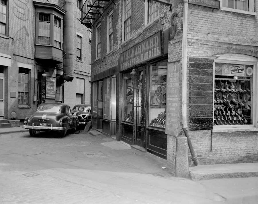 immagine in bianco e nero che raffigura un negozio di scarpe dell'epoca e delle macchine parcheggiate